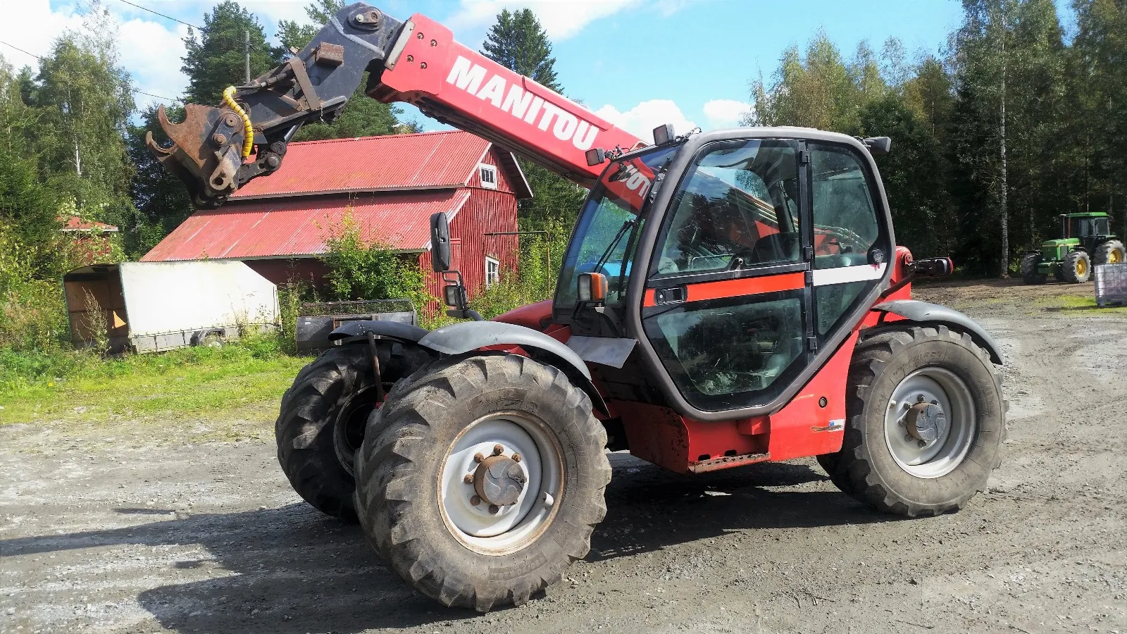 Punainen Manitou-kuormaaja pihalla maatalon edustalla, taustalla metsä ja traktori.