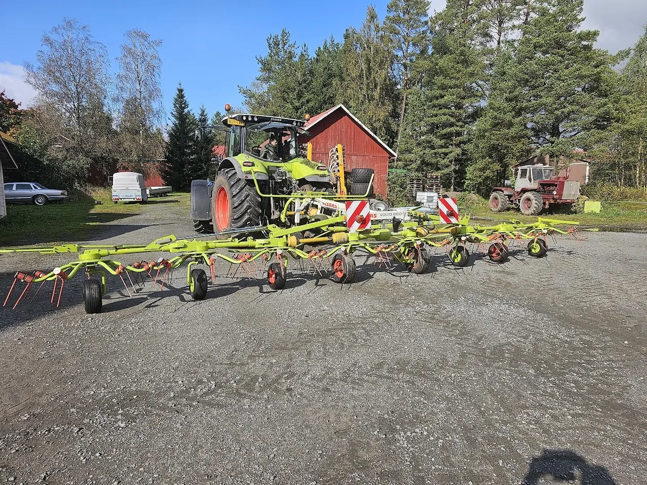 Vihreä traktori, jossa iso ja leveä keltavihreä karhotin kiinnitettynä, taustalla mökki, puita ja muita ajoneuvoja.