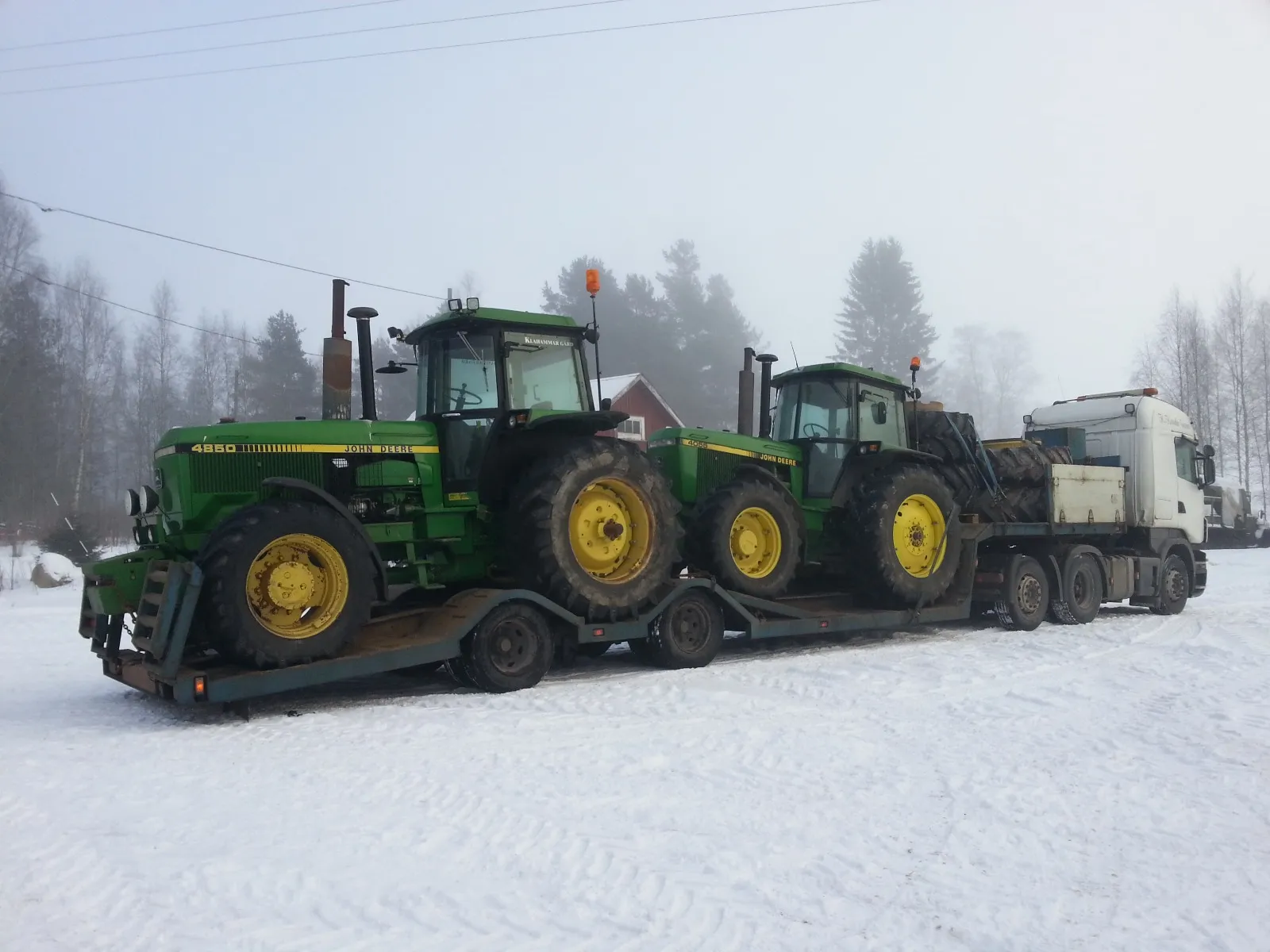 Kaksi vihreää John Deere -traktoria rekka-auton perävaunussa lumisella pihalla harmaan sumuisessa metsämaisemassa.