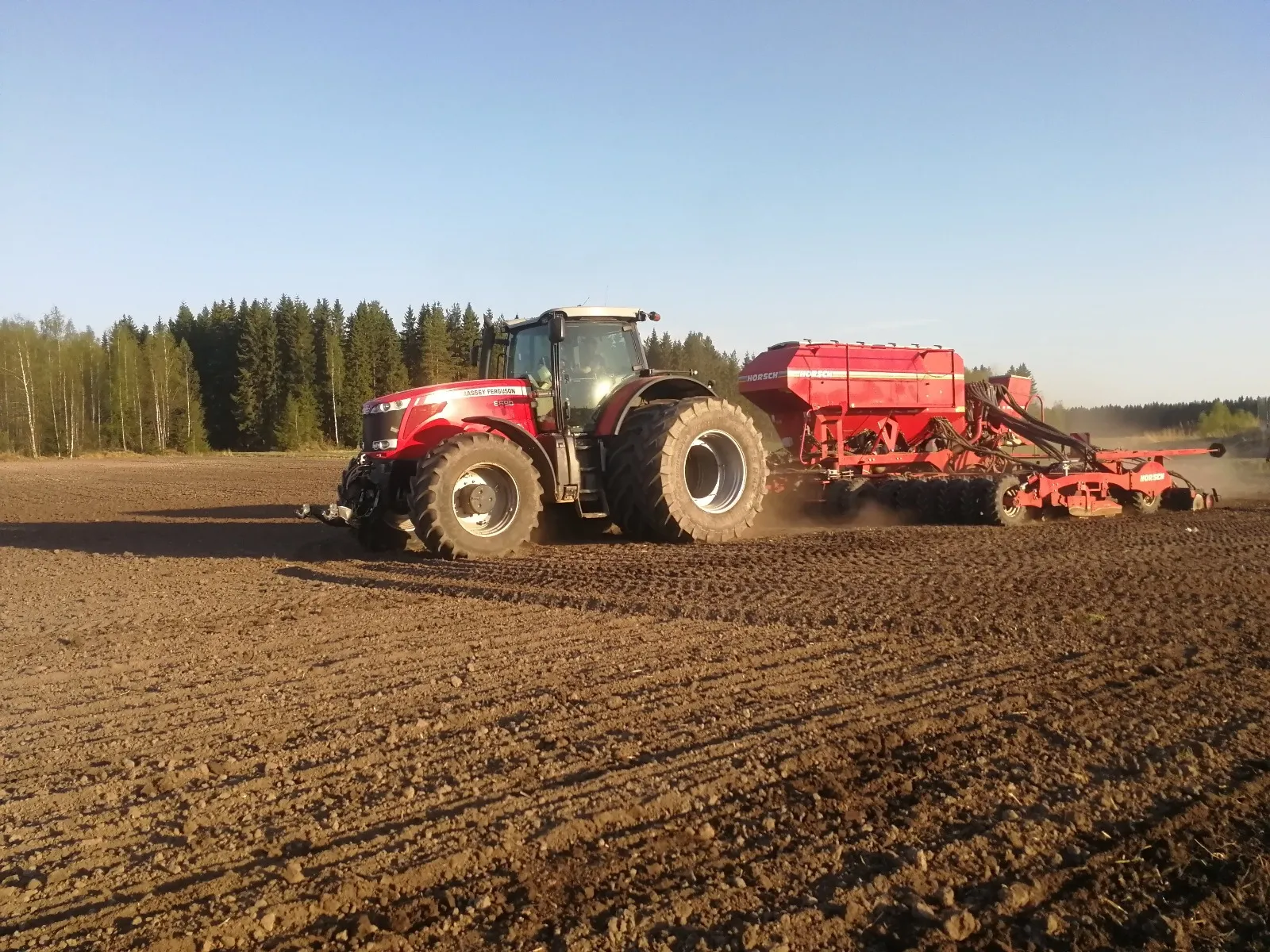 Punainen traktori vetää siemenkylvökonetta peltotöissä auringonvalossa metsätaustan edessä.