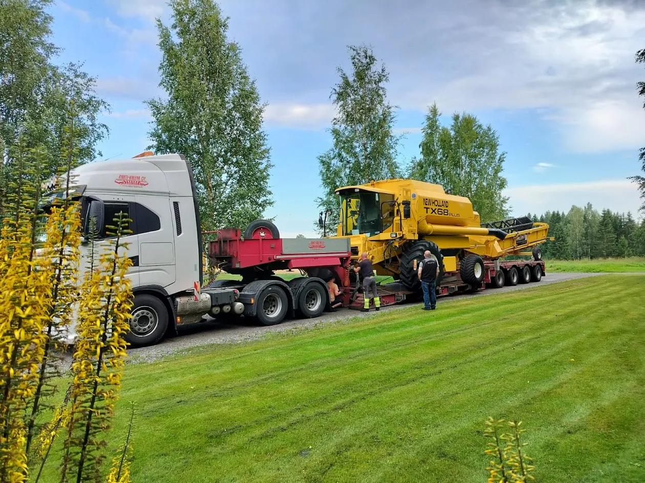 Valkoinen rekka vetää punaista lavettia, jolla on keltainen New Holland TX68 Plus -maatalouskone, ja kolme miestä työskentelee laitteen ympärillä.