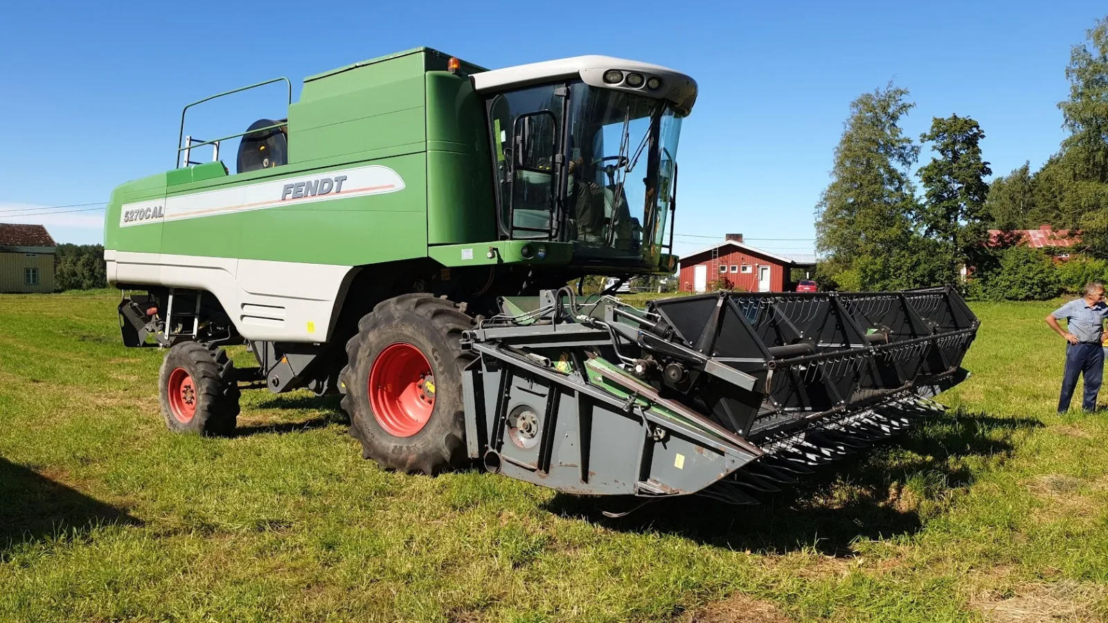 Vihreä ja valkoinen Fendt-merkkinen mallasuurna peltoaukealla sinisen taivaan alla, taustalla taloja ja puita sekä mies seisomassa oikealla.