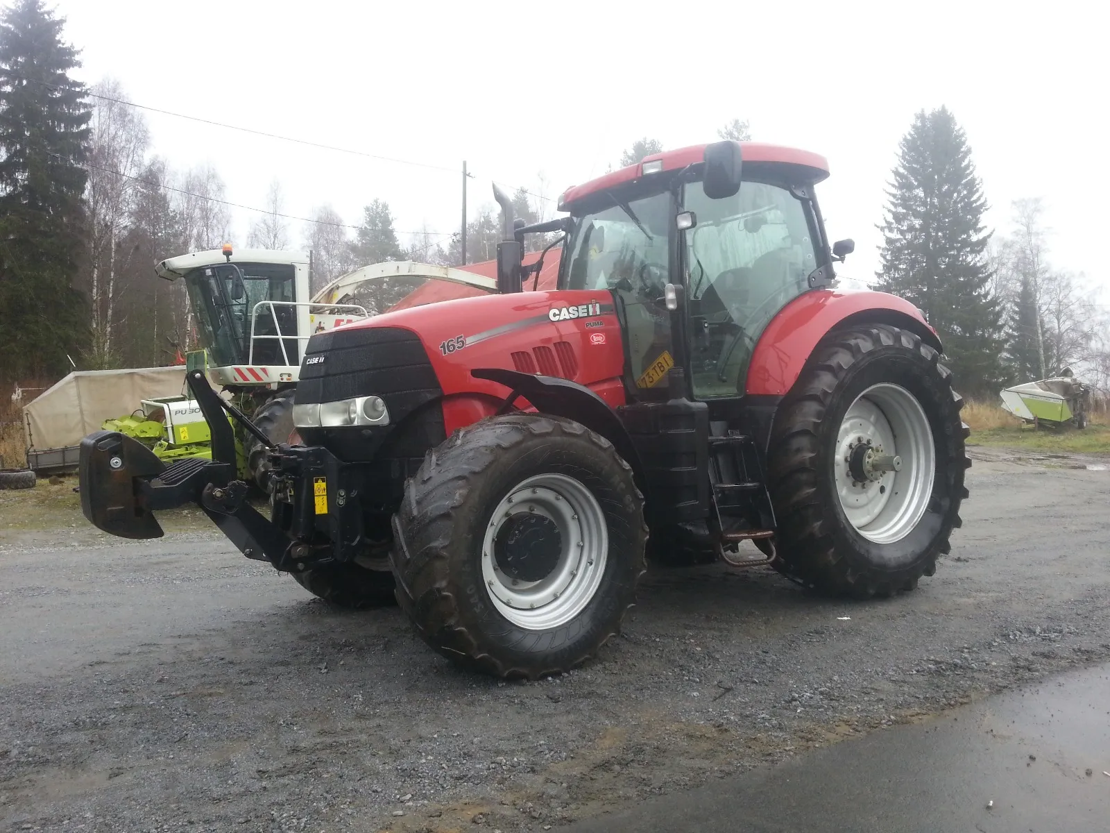 Punainen Case Puma 165 -traktori pysäköitynä soratien laitaan, taustalla metsää ja maatalouskoneita.