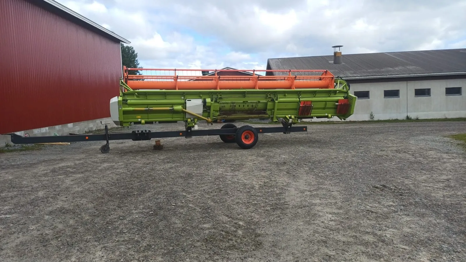 Vihreä ja oranssi maatalouskone, joka on kiinnitetty mustaan kuljetusalustaan ulkona rakennusten edessä.