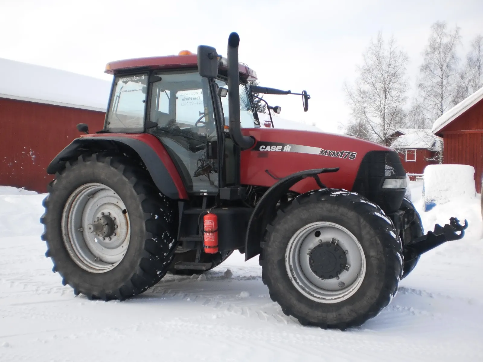 Punainen Case MXM175 traktori talvisessa maisemassa, jossa luminen maasto ja punaiset rakennukset.