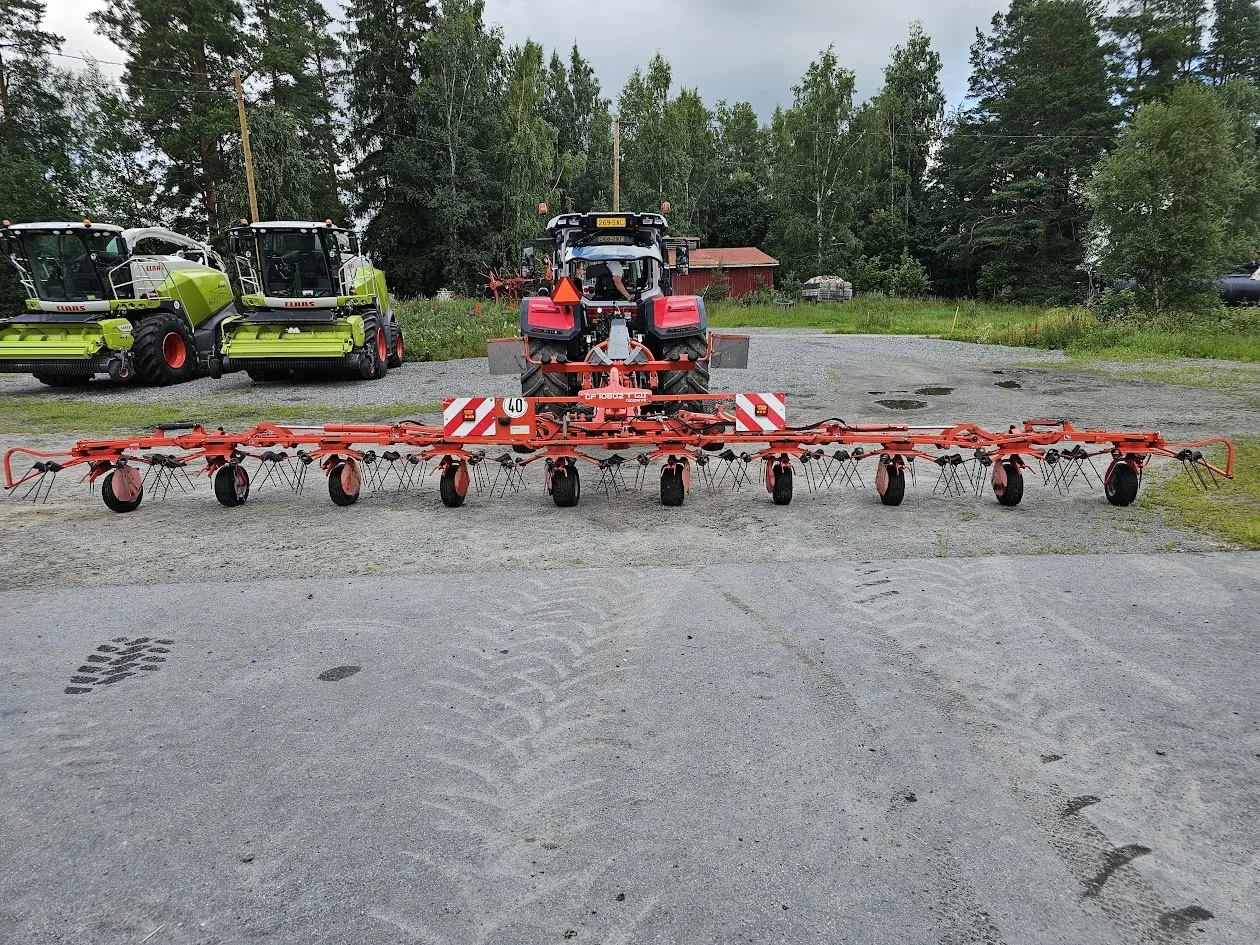 Punainen traktori, johon kiinnitetty laaja karhotin, pysäköity sorapihalle metsän edustalla.
