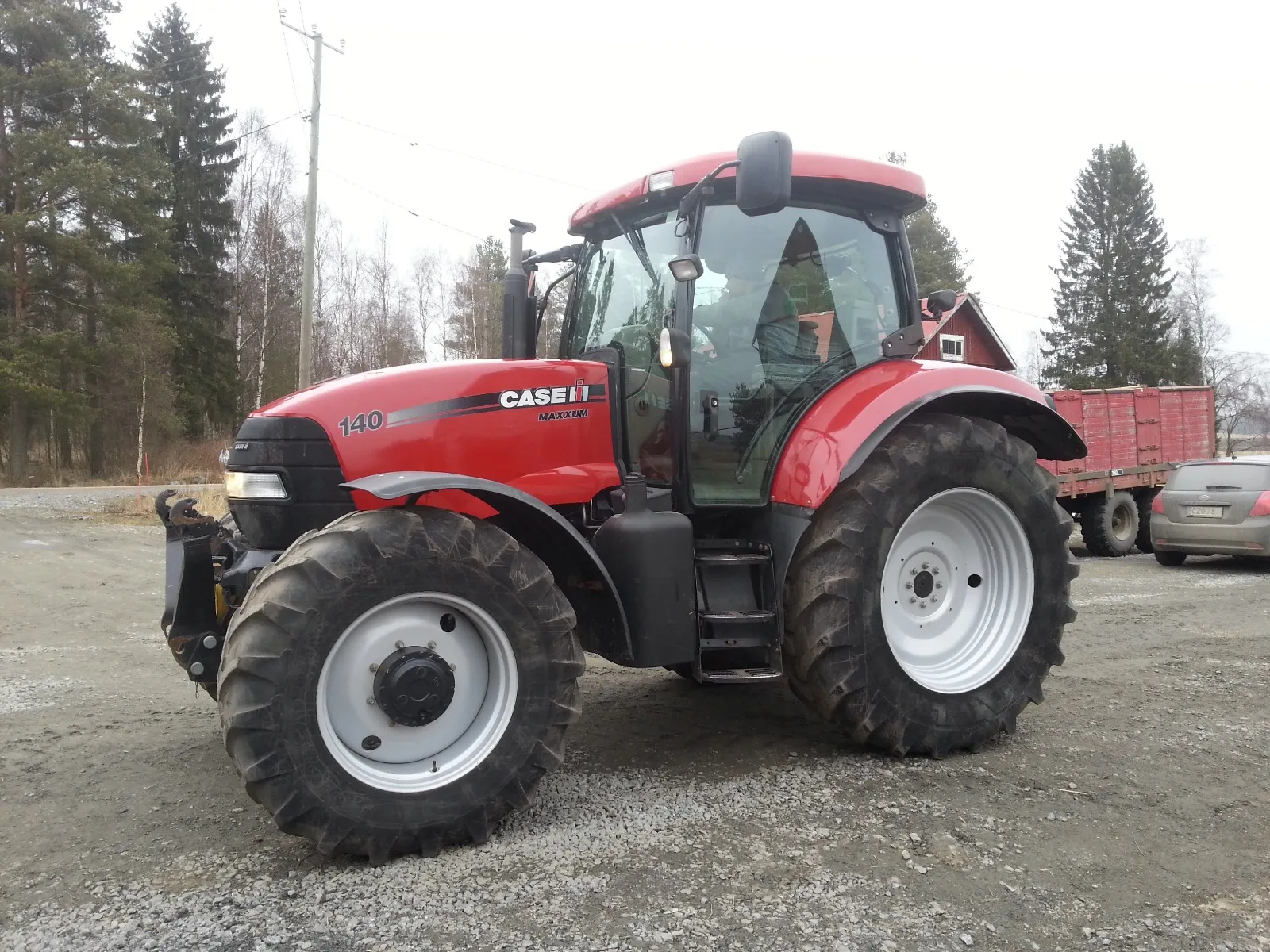 Punainen CASE 140 Maxxum -traktori sorapintaisella tiellä pihalla, taustalla kuorma-auto ja auto.