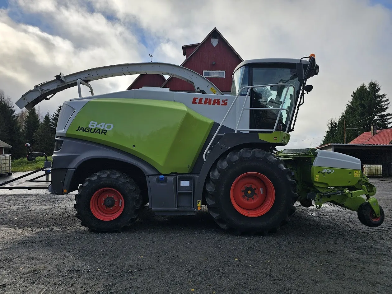 Vihreä ja harmaa CLAAS Jaguar 840 -rehuntekniikkakone maassa, taustalla punainen rakennus ja pilvinen taivas.
