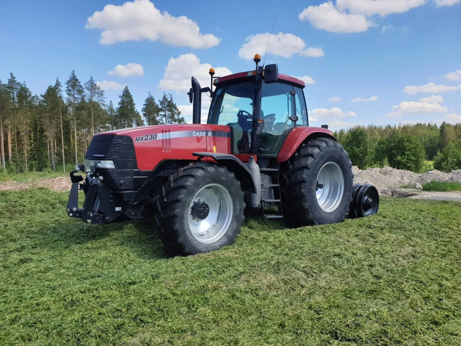 Punainen Case MX230 -traktori vihreällä nurmella metsätaustan edessä sinisen taivaan alla.