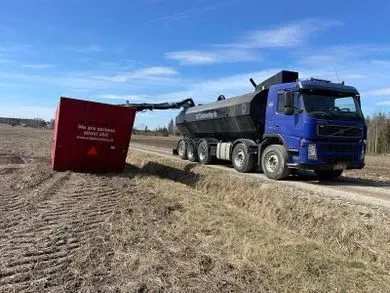 Sininen kuorma-auto lastaa multaa tai kompostia maassa olevaan punaisen astian säiliöön avoimella pellolla.