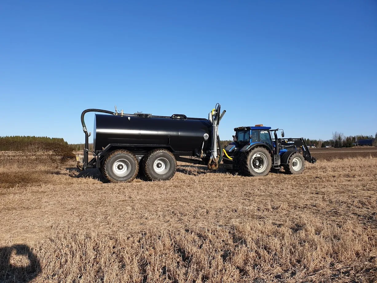 Sininen traktori kytkettynä mustaan lietteenkuljetussäiliöön kuivalla pellolla kirkkaana päivänä.