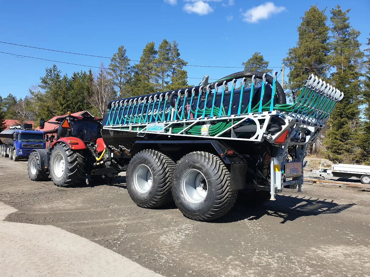 Punainen traktori vetää suurta mustaa nestesäiliötä, jossa on vihreitä letkuja ja metallinen järjestelmä, taustalla puita ja sininen taivas.