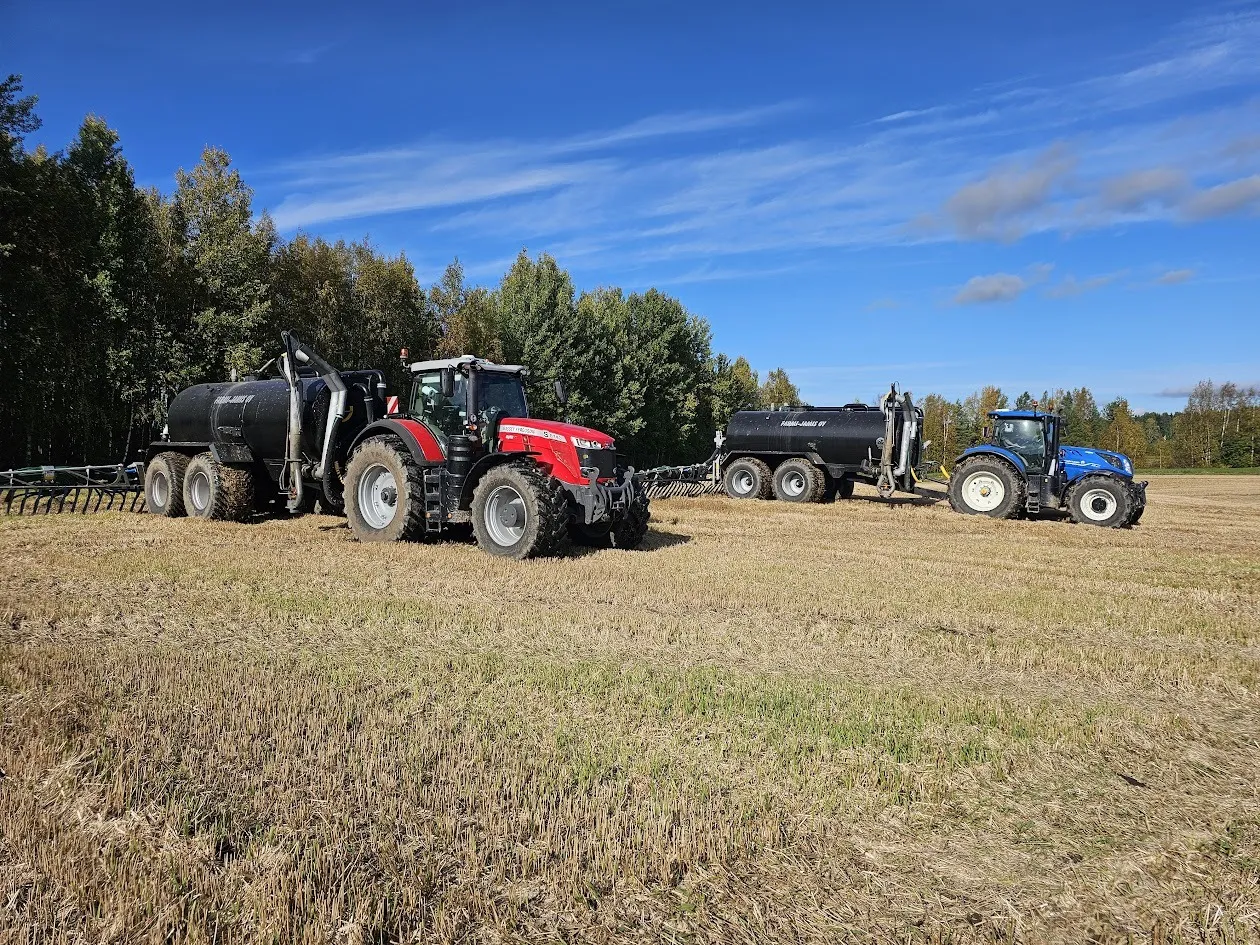 Kaksi traktoria, joissa kummassakin musta lietevaunu, pellolla sinisen taivaan alla ja metsän reunassa.