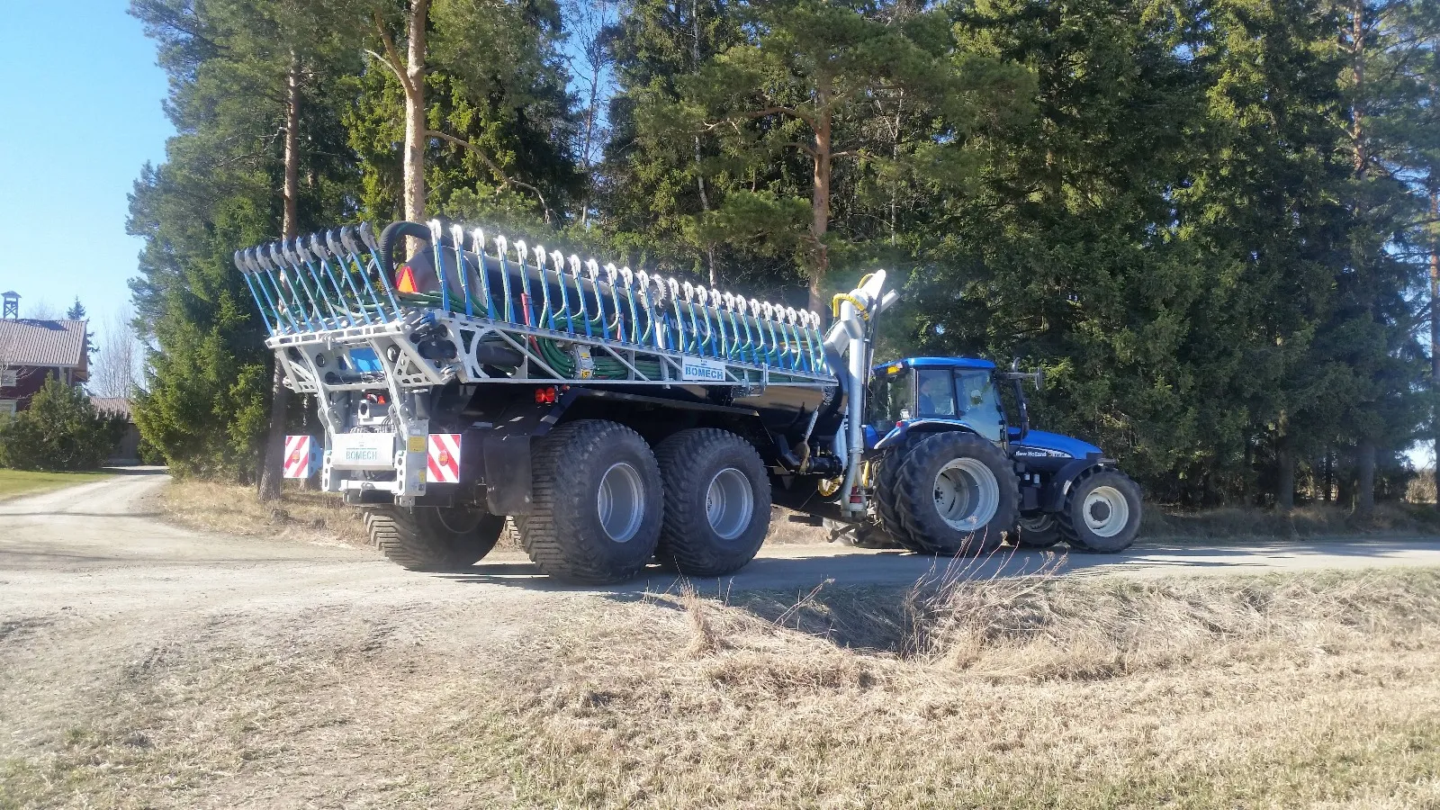 Sininen traktori on kiinni suurikokoisessa ja monirengasaisessa maatalousperävaunussa, joka on varustettu putkilla ja letkuilla, ja kuvassa on taustalla metsä ja maatila.