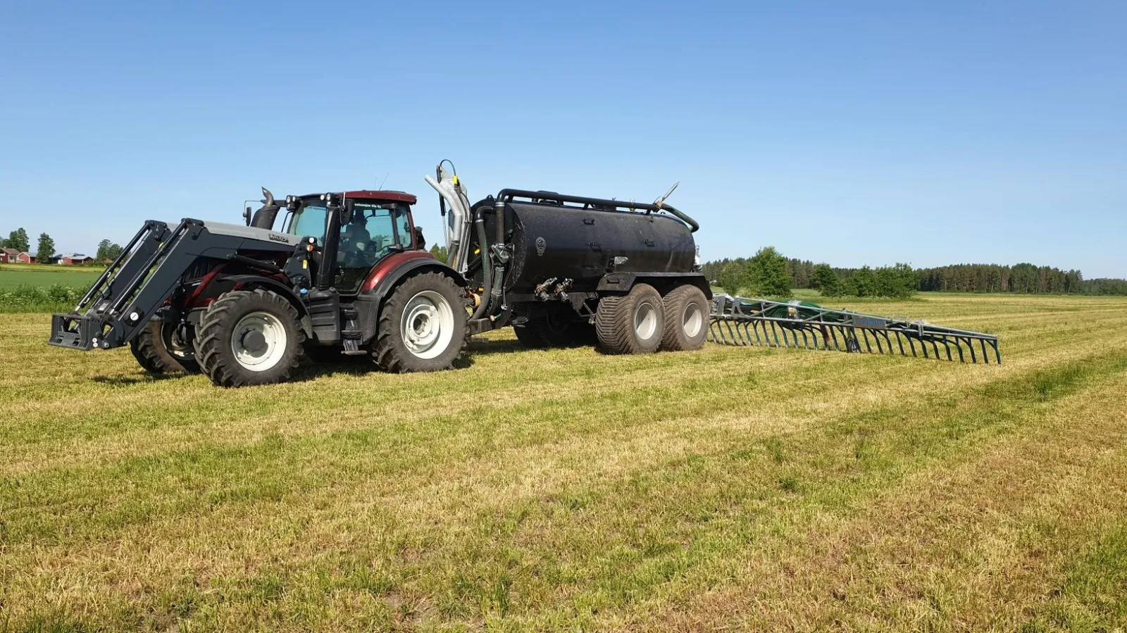 Punainen traktori, joka vetää mustaa lietekärryä pellolla sinisen taivaan alla.