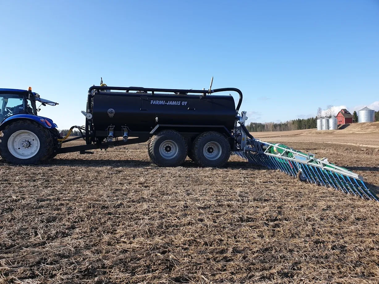Traktori vetää mustaa lietteenkuljetusvaunua pellolla kirkkaan sinisen taivaan alla.
