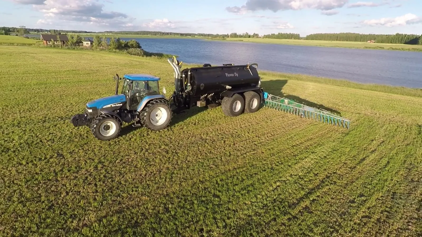 Sininen traktori vetää mustaa lannoitteen levityssäiliötä kuivalle pellolle järven rannalla.