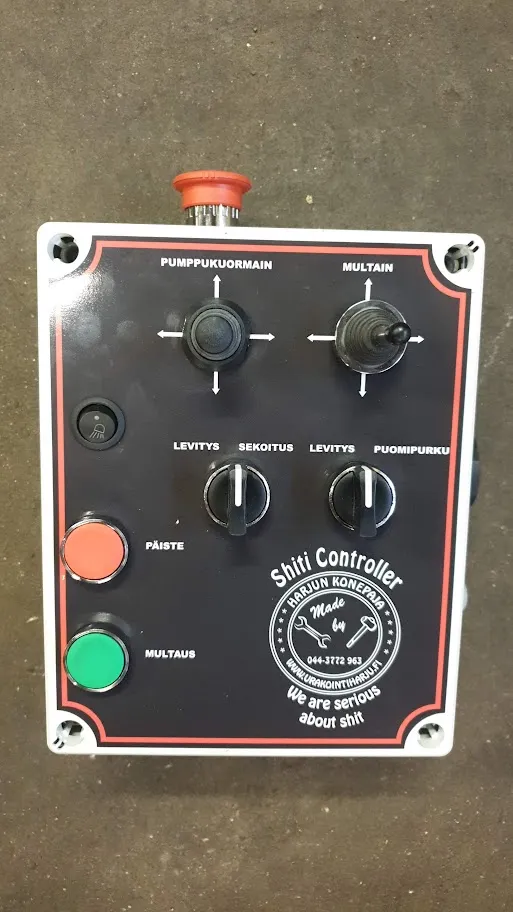 Suomenkielinen teollisuusohjainpaneeli, jossa kaksi joystickia, kaksi mustaa säätönuppia ja kolme värillistä painiketta betonipinnalla.