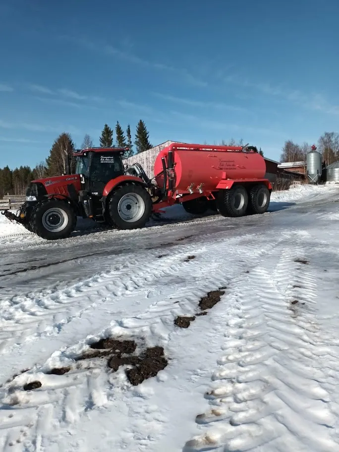 Punainen traktori, joka vetää isoa punaista nestettä sisältävää säiliövaunua lumisella maatilalla.