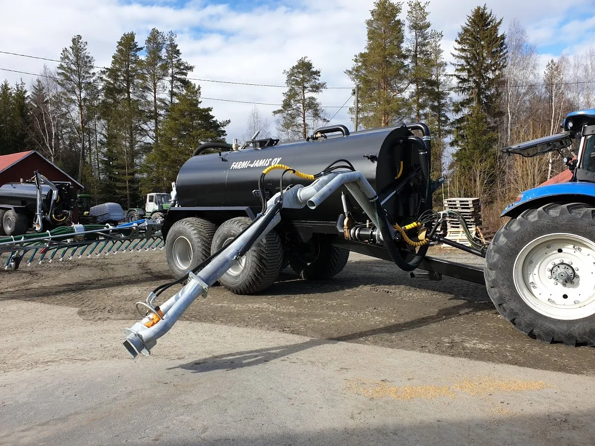 Musta lietekärry kiinnitetty siniseen traktoriin, ympärillä puita ja rakennuksia.