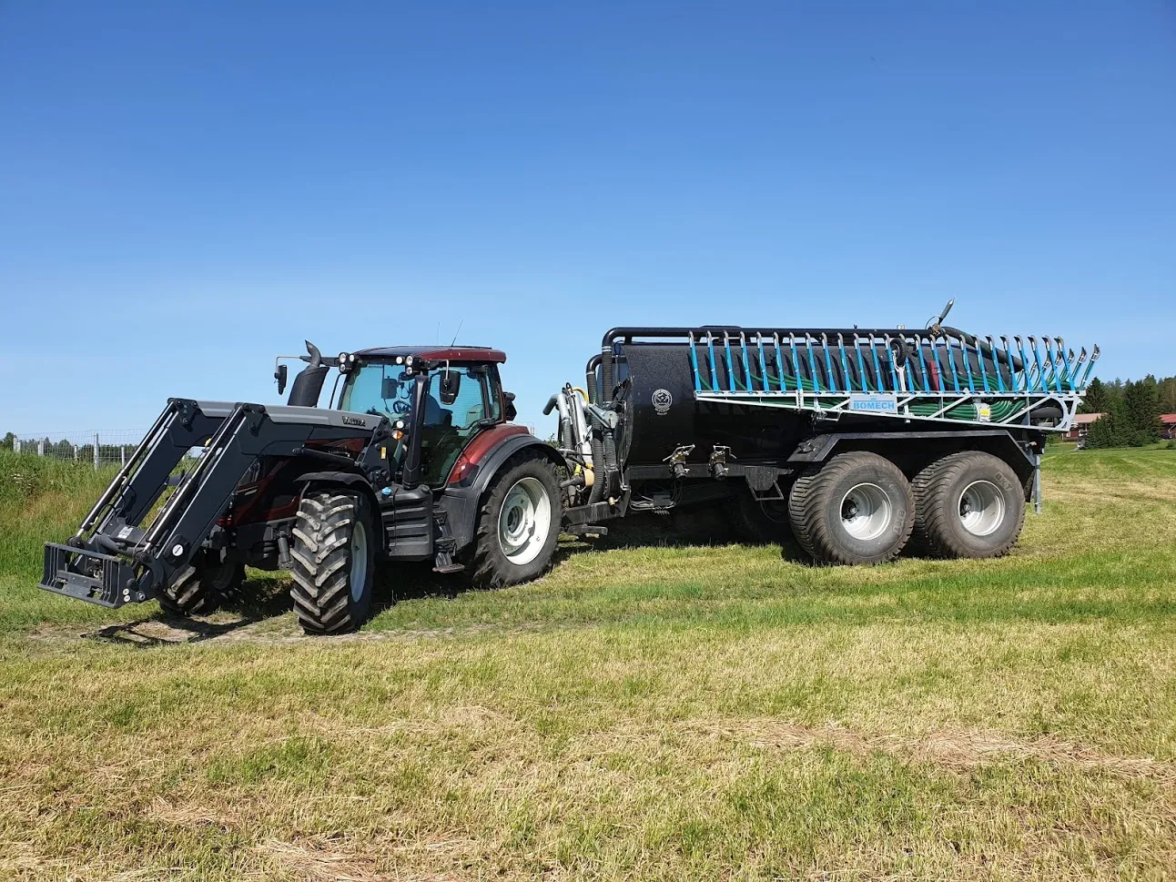 Punainen traktori kiinnitettynä mustaan lietevaunuun nurmikolla kirkkaana päivänä.