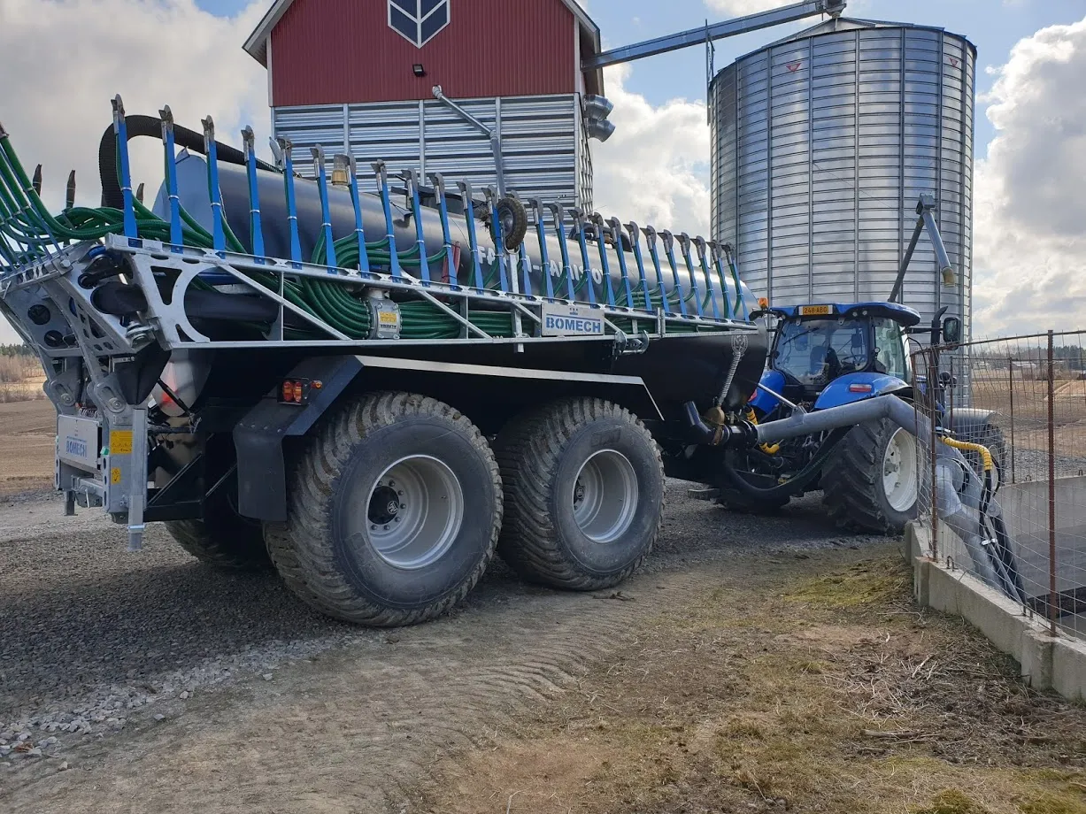 Maatalouskone, jossa Bomech-merkkinen puoliperävaunu ja sininen traktori pihalla lähellä viljasäiliötä ja latoja.