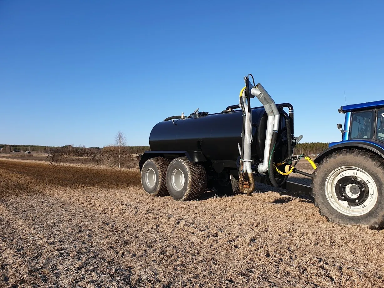 Musta lietelietevaunun ja sinisen traktorin yhdistelmä pellolla kirkkaana päivänä.