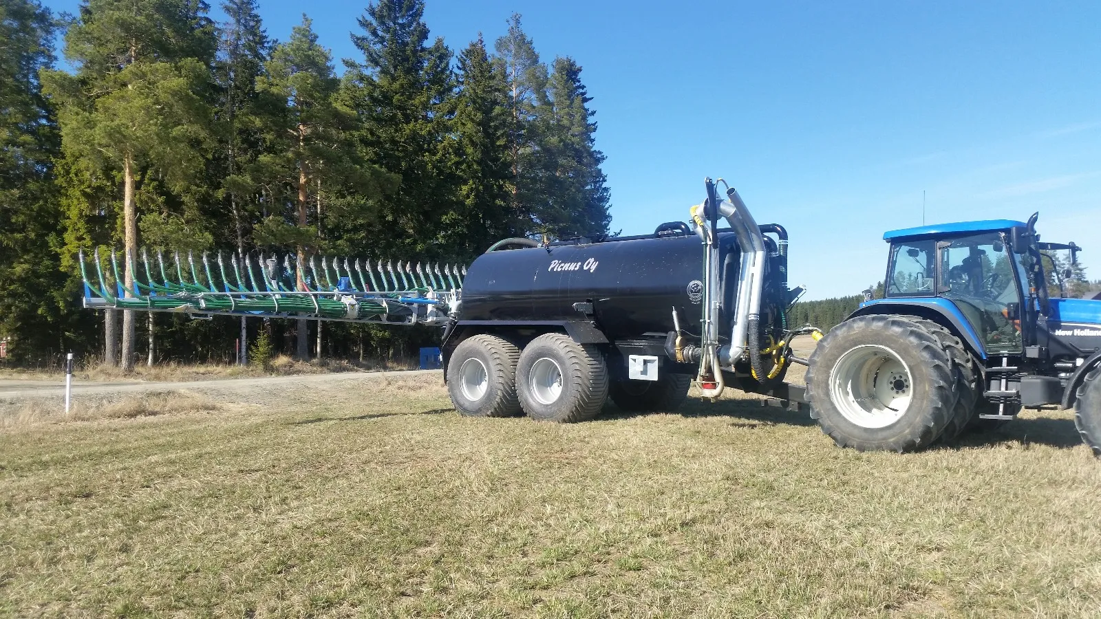 Sininen traktori vetää mustaa lietelannan levityskärryä avoimella nurmialueella, taustalla metsä ja sininen taivas.
