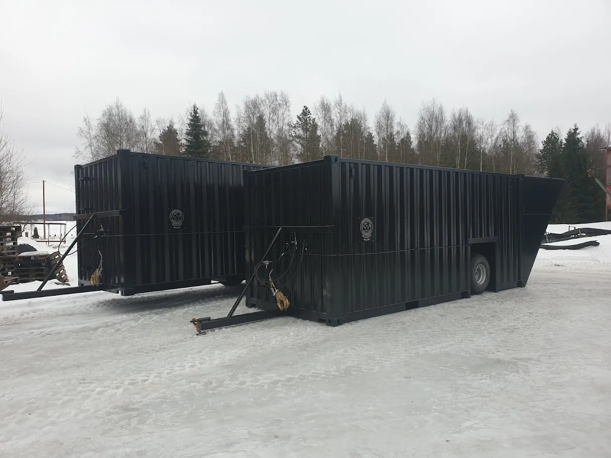 Kaksi mustaa jätelavaa talvisessa ulkoilmassa lumisella maalla, taustalla voivat näkyä puut ja hämärä taivas.