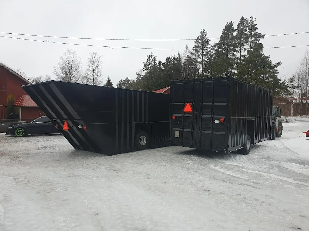 Kaksi mustaa jätelaatikkoa lumisella pihalla talojen ja mäntyjen edessä.