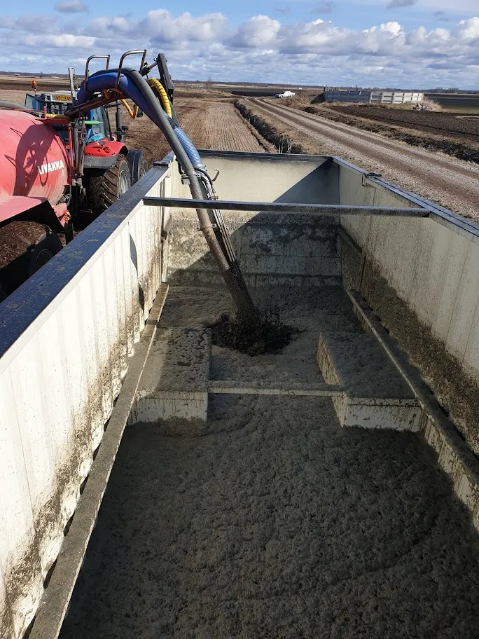 Traktori pumppaamassa lietettä suureen rekkaan maaseutumaastossa pilvisenä päivänä.