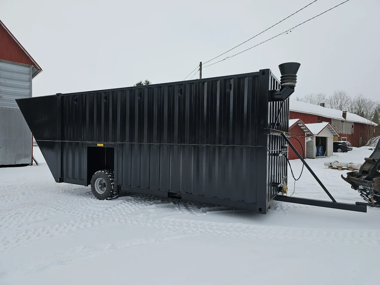 Musta, suuri siirrettävä lietekontti lumisella pihalla punaisen navetan edessä.