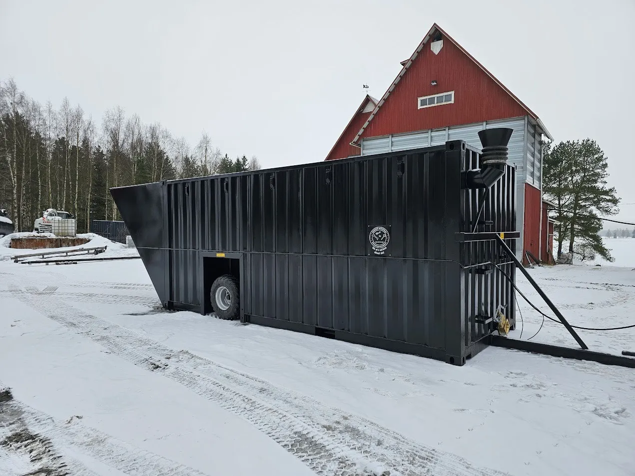 Mustan metallisen lietekontin ulkonäkö lumisessa pihassa punaisen rakennuksen vieressä.