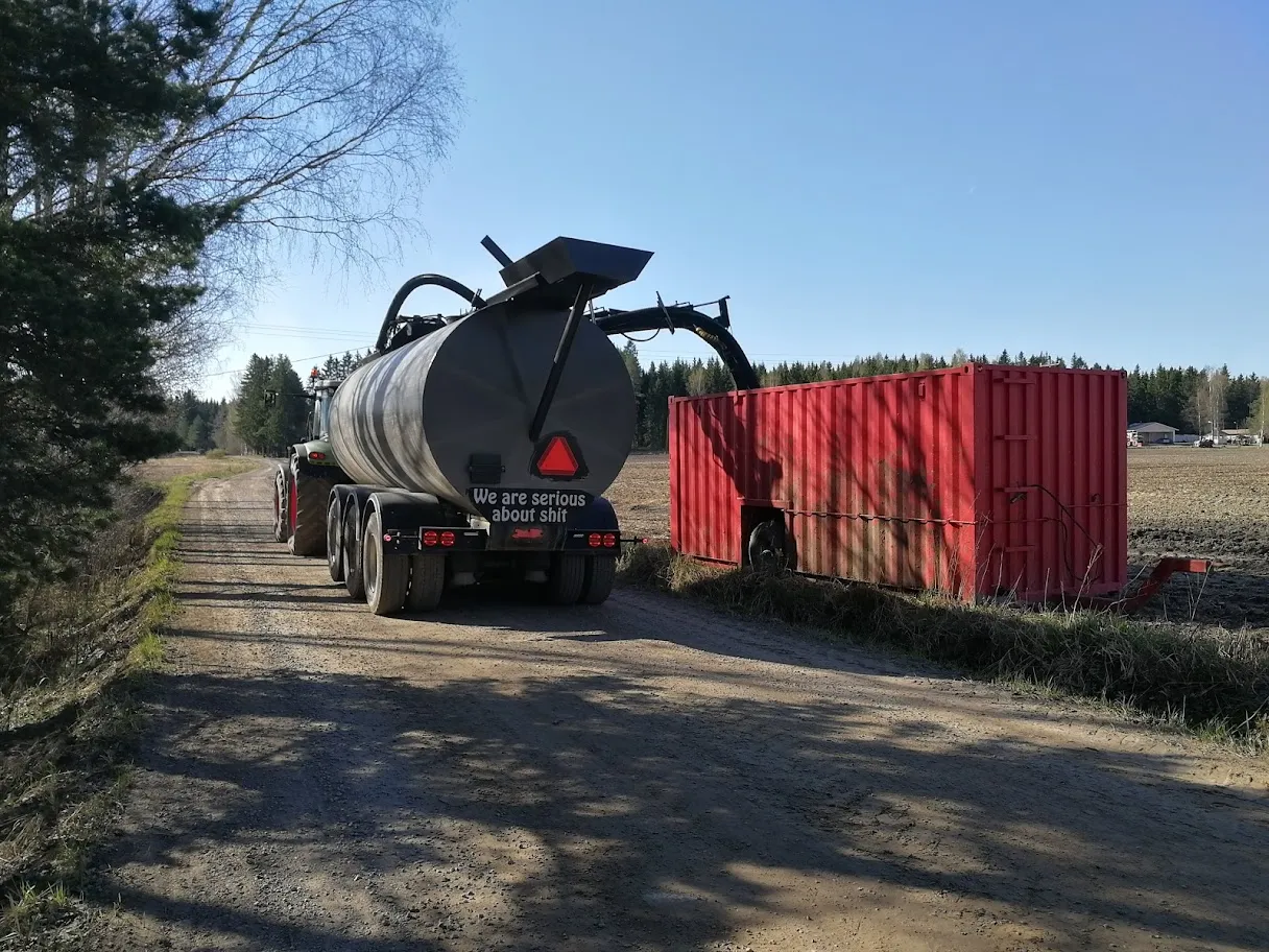 Maatilalla hiekkatiellä oleva renkavaunu ja suuri punainen kontti, taustalla peltoa ja metsää.