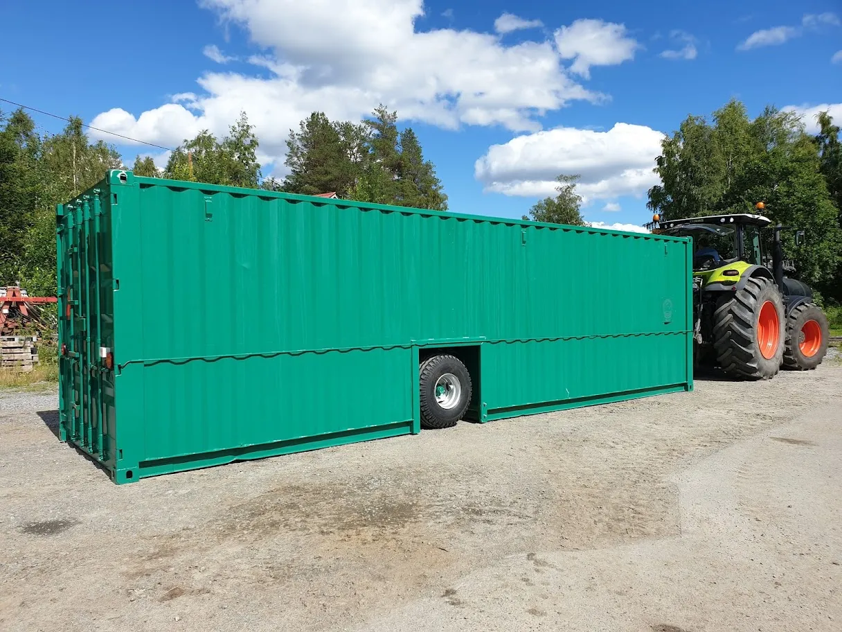 Vihreä säiliökontti, jonka alareunassa on aukko pyörälle, peräkärrynä traktorin edessä metsämaisemassa.