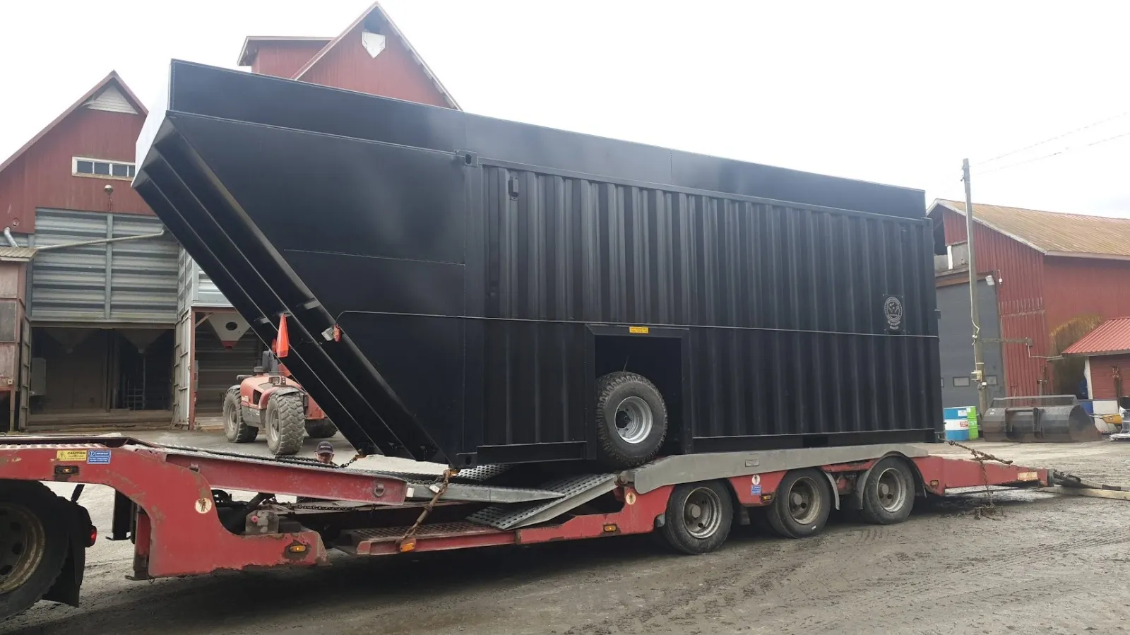 Musta suuri lietekontti traktorin ja maatalousrakennusten edessä kiinnitettynä punaisen perävaunun kyytiin.