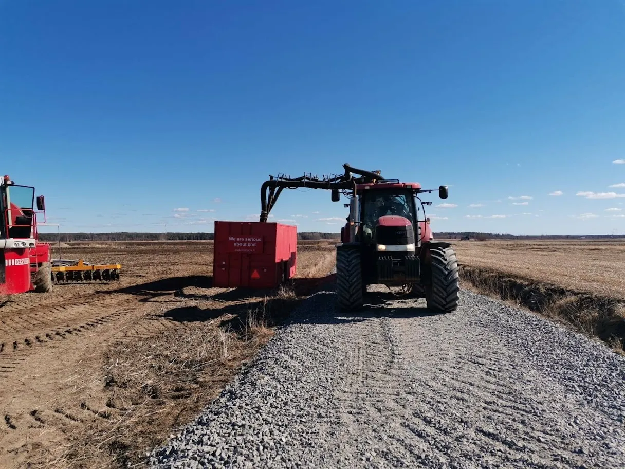 Punainen traktori kippikärryllä kiviaitauksella pellon laidalla sinisen taivaan alla.