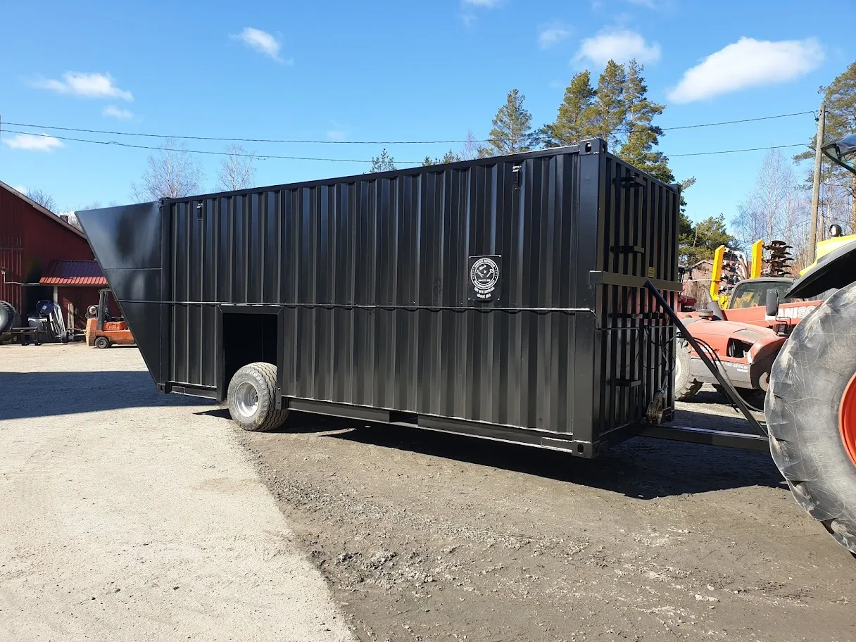 Musta metallinen lietekontti pyörän päällä, vedettynä traktorilla pihamaalla.