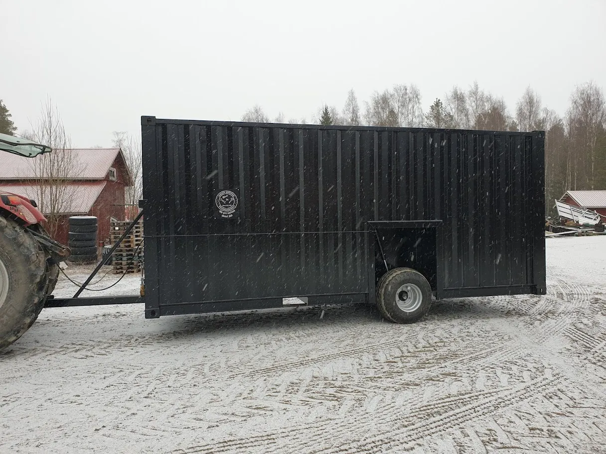 Musta metallinen lietekontti, jota vetää punainen traktori lumisella pihalla harmaana talvipäivänä.