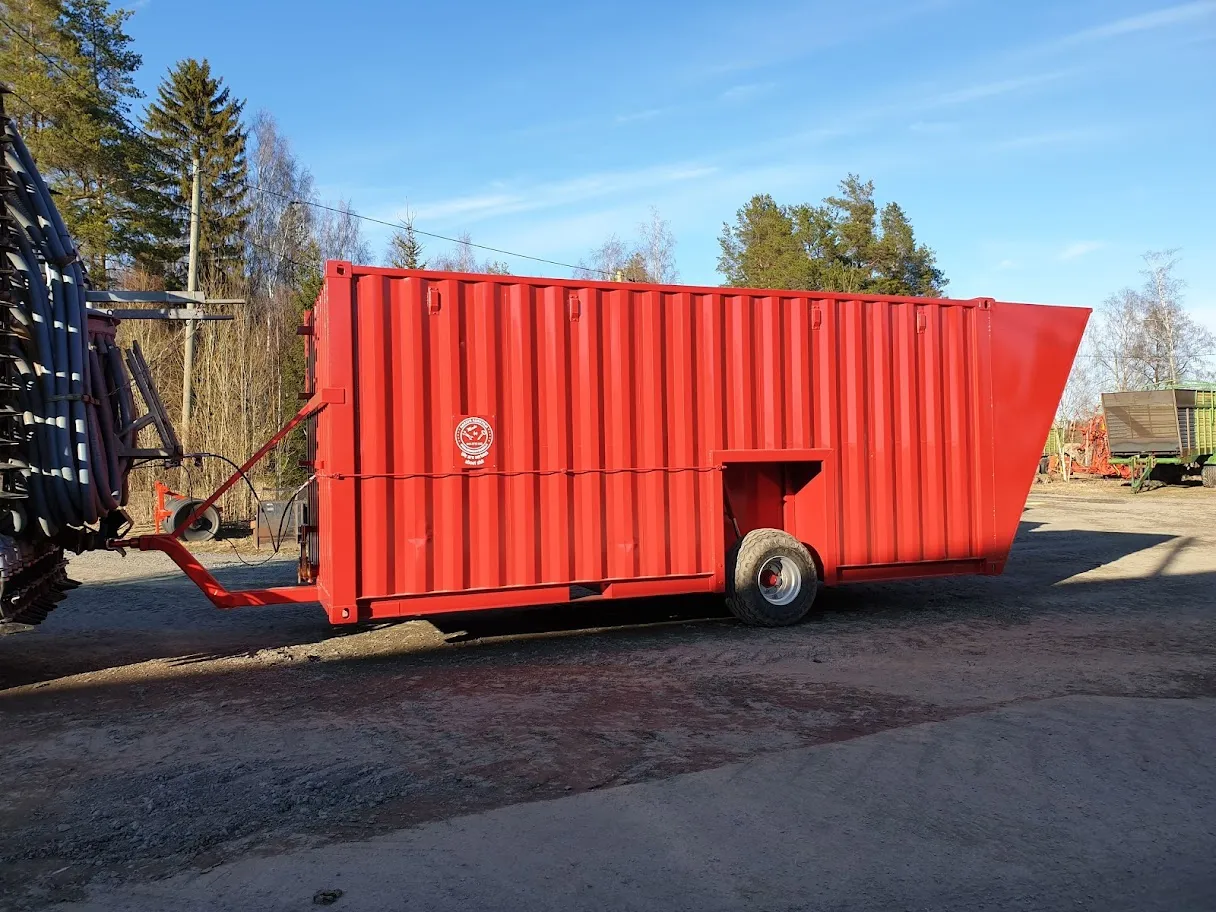 Punainen lietelantakontti maassa, kiinnitetty traktoriin, taustalla puita ja selkeä sininen taivas.