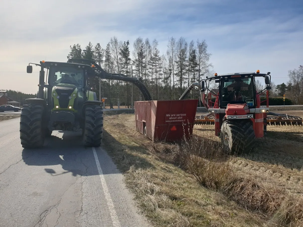 Kaksi traktoria maantie- ja peltomaisemassa, joista toinen siirtää leikettyä satoa punaisella perävaunulla varustettuun säiliöön.
