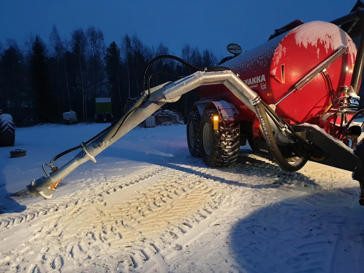 Punainen lietesäiliövaunu ja pitkä putkimainen torvi lumisella pihamaalla talvi-illan hämärässä.