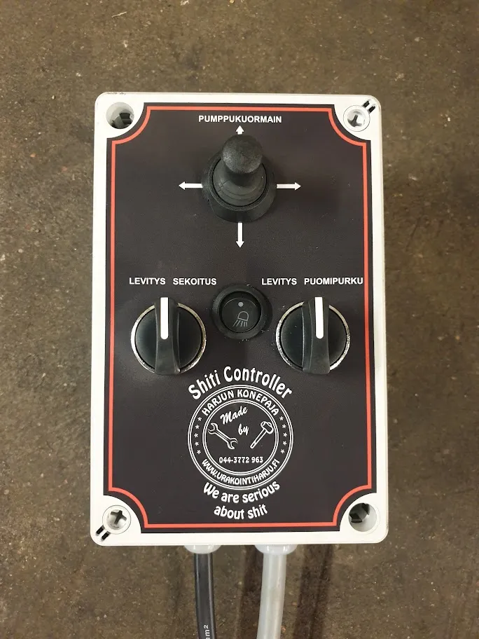 Ohjauspaneeli, jossa joystick ja kaksi säätönuppia sekä tekstiä suomeksi ja englanniksi, mukana käännös "We are serious about shit".