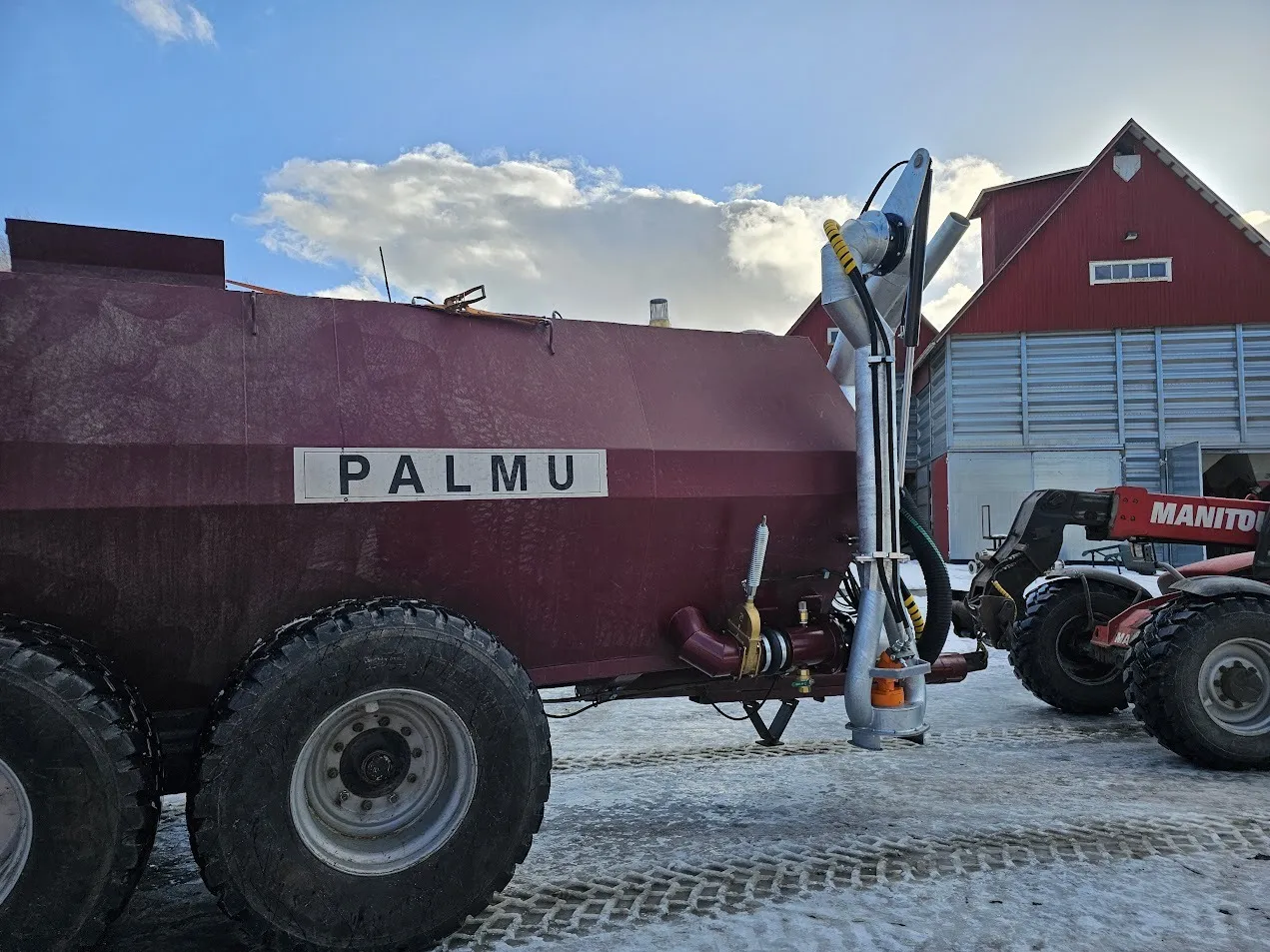 Punainen PALMU-merkitty peltovaunu ja Manitou-kuormain lumisella piha-alueella maatilan rakennusten edessä.