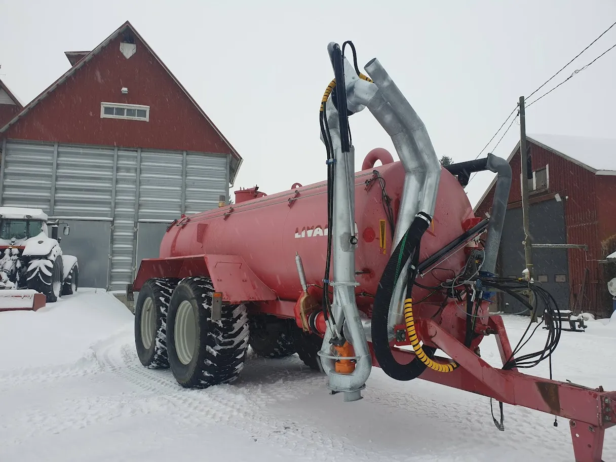 Punainen maatalouskone, jossa säiliö ja putkisto, lumisessa pihassa punaisen navetan edessä.