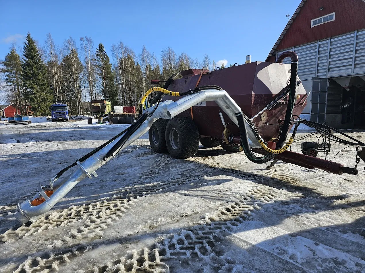 Maatalouskone isoilla renkailla, varustettuna pitemmällä metalliputkella lumisella pihamaalla maatilan rakennuksen vieressä.