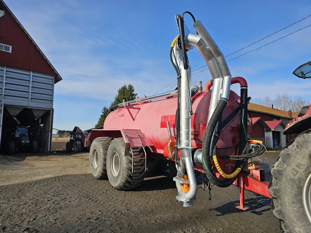 Punainen lietevaunu ja traktori maatilalla kirkkaana päivänä.