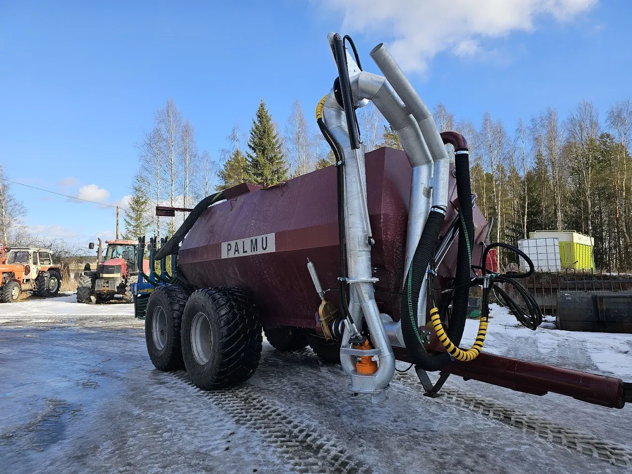 Punainen Palmu-merkkinen lietelannan levitykseen tarkoitettu maatalouskone jään peittämällä pihamaalla, taustalla traktoreita ja puiden rivi.