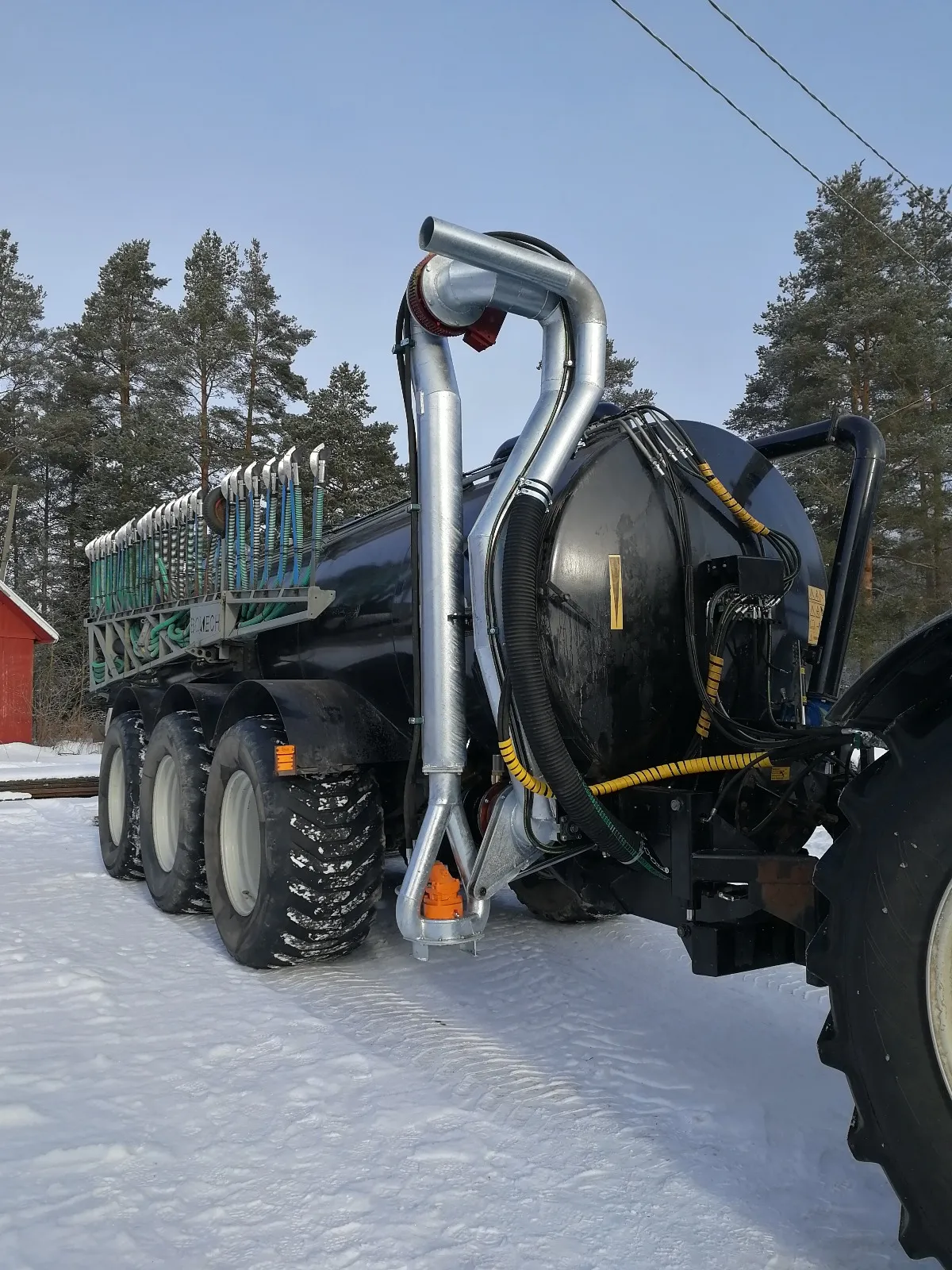Musta järeä lietevaunu traktorin kiinnityksessä lumisella pihamaalla, taustalla mäntyjä ja punainen rakennus.