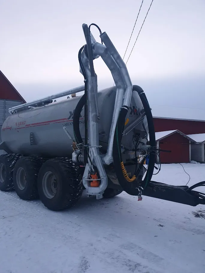 Maatalouskone, jossa suuri metallitankilla varustettu tankki neljällä suurella renkaalla, talvimaisemassa lumisella pihalla.